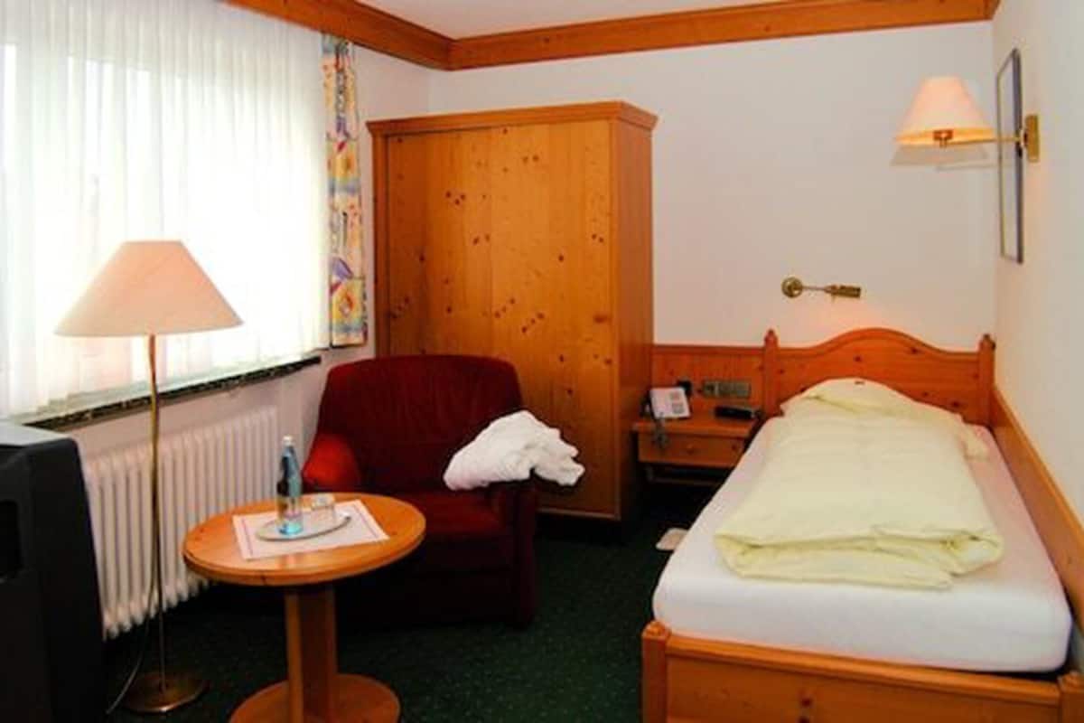 A hotel room with a single bed, a red armchair, a small round table with a lamp, and a wooden wardrobe.