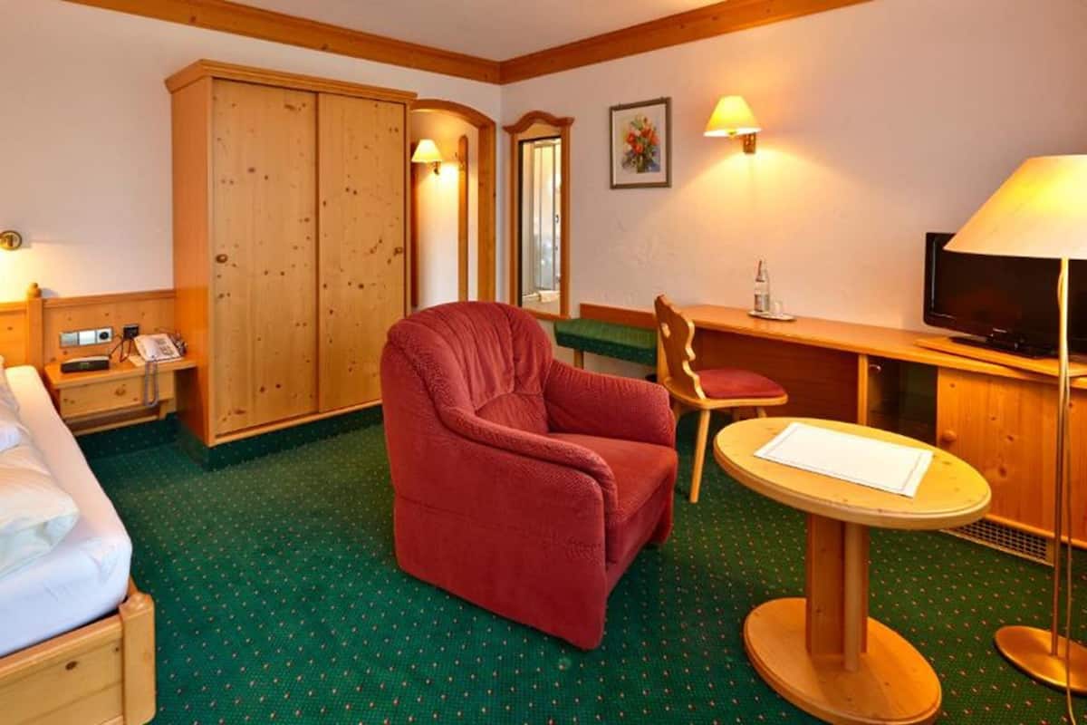 A hotel room with a bed, a red armchair, a small desk with a chair, a television, and a wardrobe.