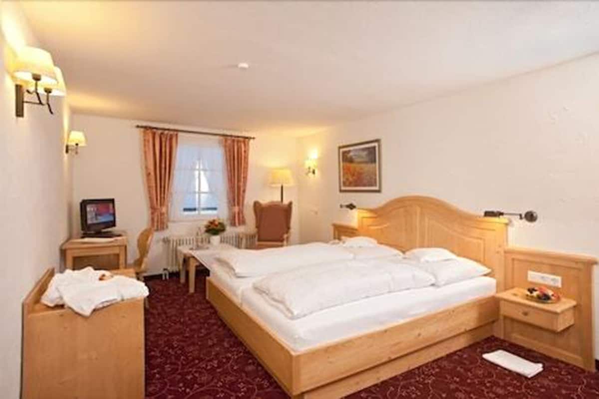 A hotel room with a large bed, a desk with a computer, a chair, and a small table with a lamp.