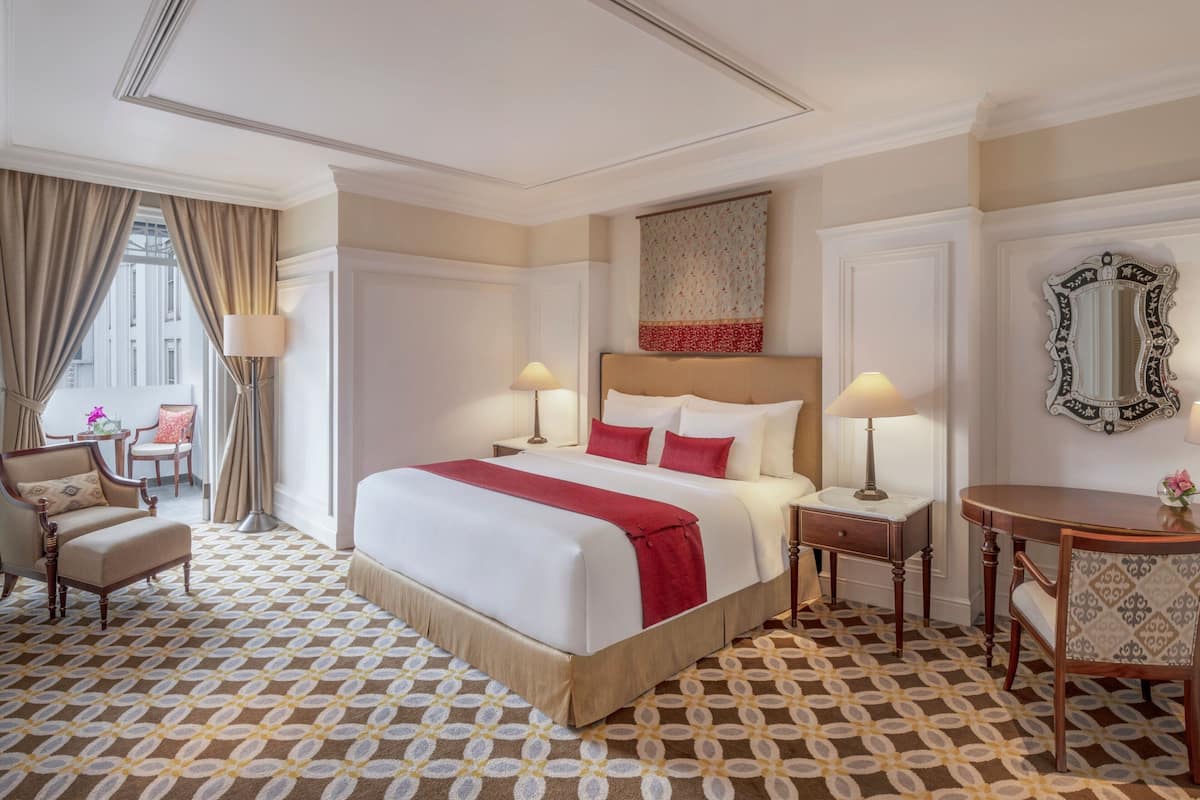 A hotel room with a large bed, two bedside tables, a chair, a small table, and a mirror. The room has patterned carpet and curtains.