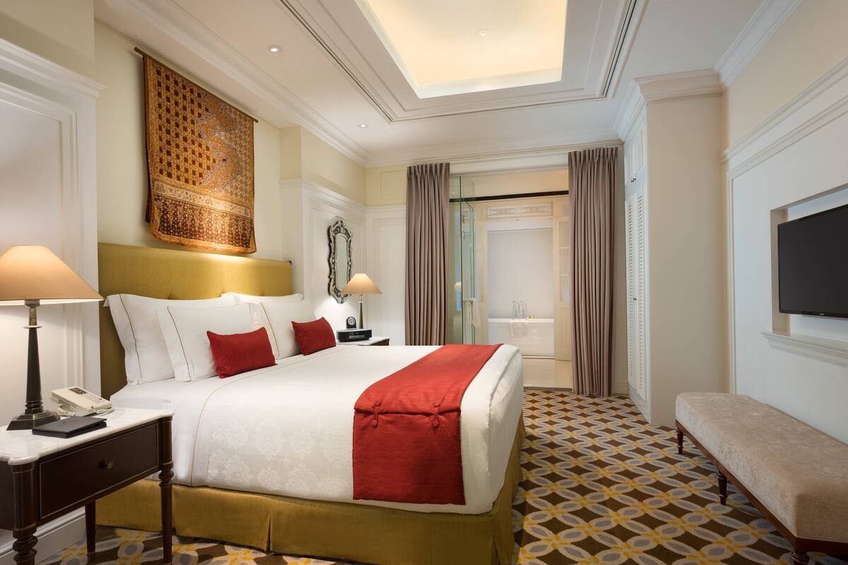 A hotel room with a large bed, bedside tables, a bench, a TV, and a bathroom visible through a glass door.