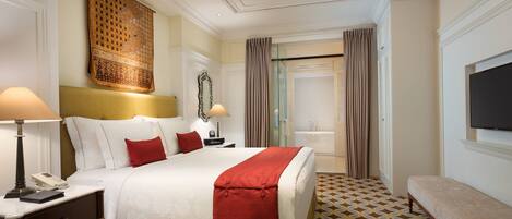 A hotel room with a large bed, bedside tables, a bench, a TV, and a bathroom visible through a glass door.