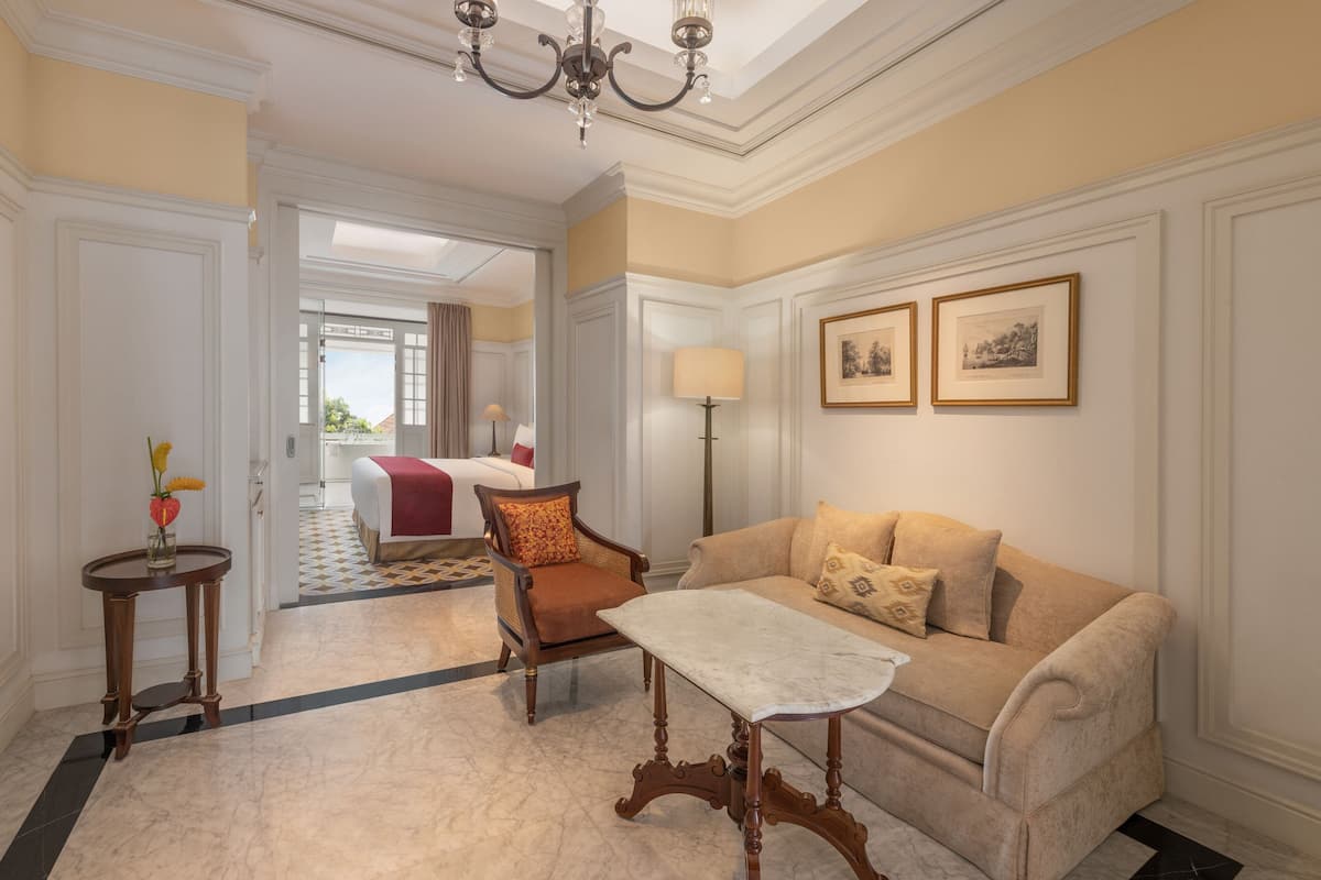 A hotel room with a beige sofa, a wooden chair, a small table, and a lamp. There are two framed pictures on the wall.