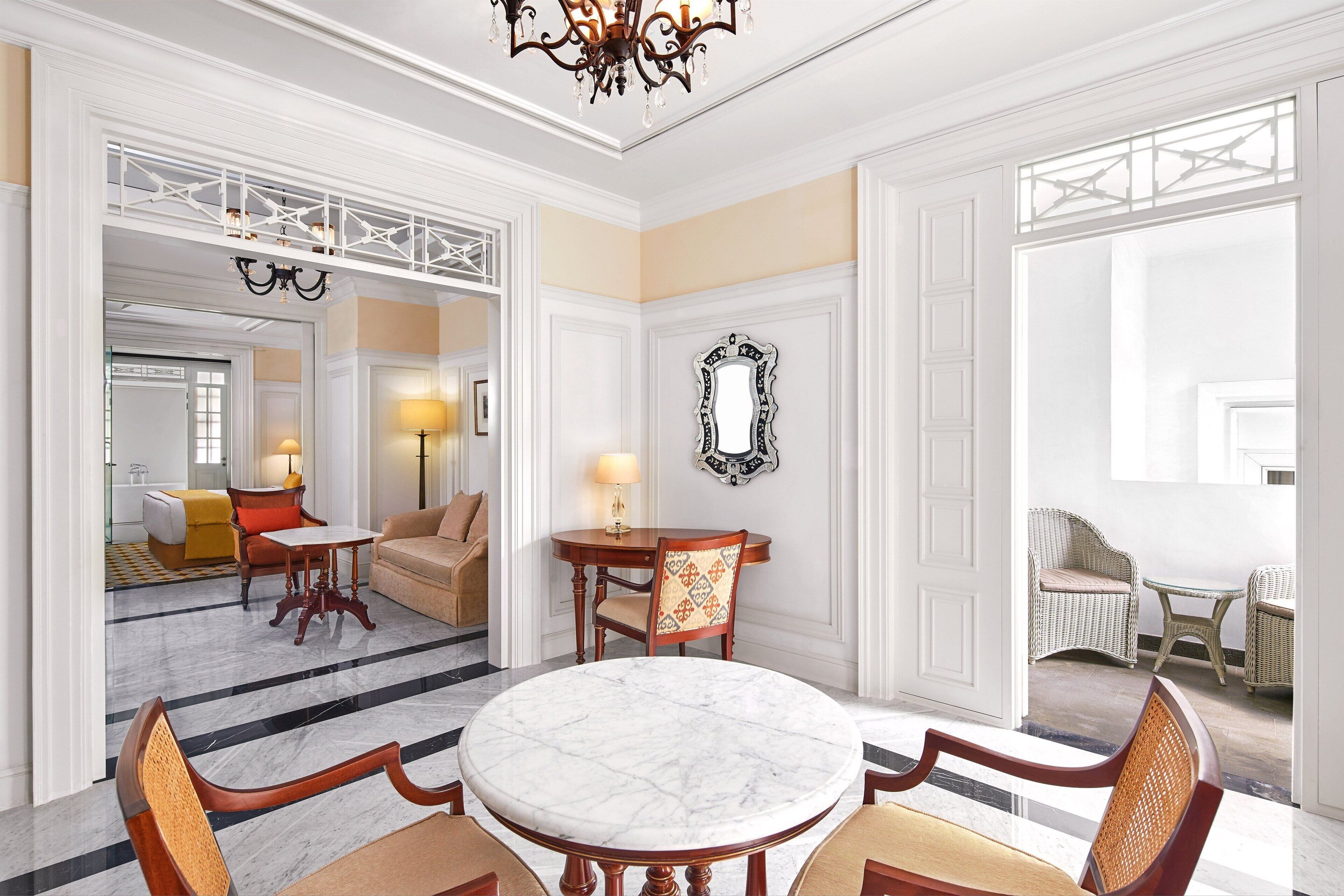 A spacious room with a marble floor, a round table with chairs, a sofa, and a dining table with chairs. There is a chandelier, a mirror, and a view of a balcony with wicker furniture.