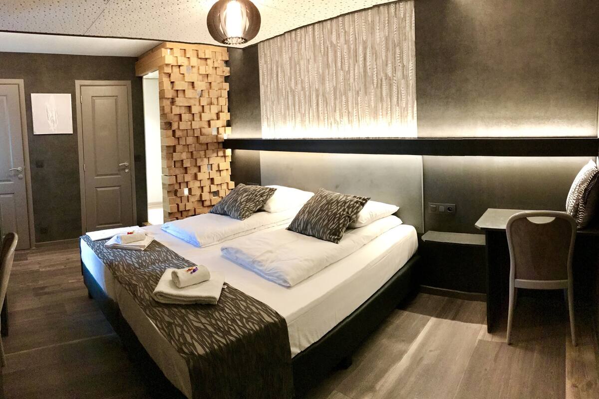 A modern hotel room with a large bed, a wooden headboard, a desk, and a chair.