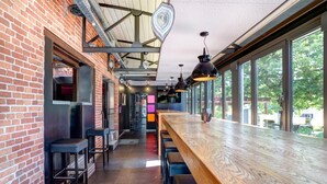 A modern bar with a long wooden counter, black bar stools, and large windows offering a view of the outdoors. - RAVEL Hotel Cyrano (Waimes)