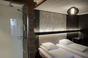 A modern bathroom with a shower, a bed with white linens, and a desk with a chair. - RAVEL Hotel Cyrano (Waimes)