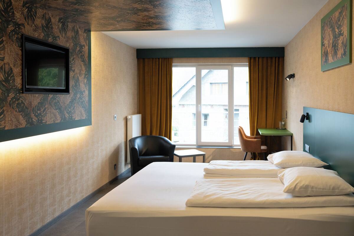 A hotel room with a bed, a chair, a small table, and a window with curtains.