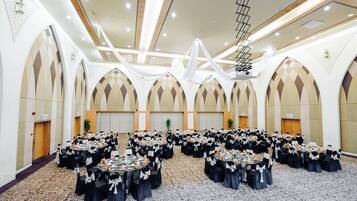 A large banquet hall with multiple round tables set up for a formal event, featuring a high ceiling with a central light fixture and arched wall designs.