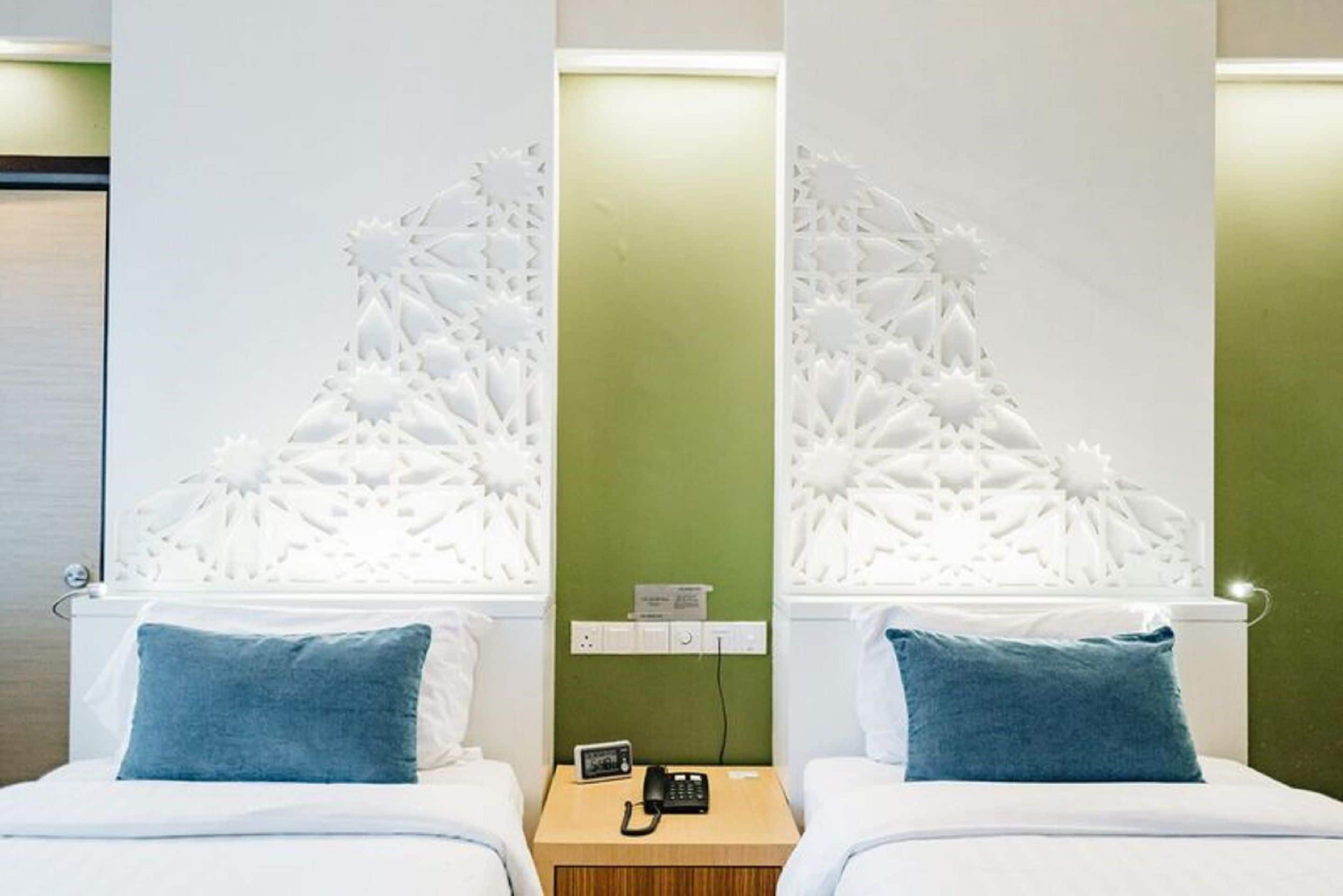 Ein Hotelzimmer mit zwei Betten, jeweils mit einem blauen Kopfkissen, einem Nachttisch aus Holz mit Telefon und einem kleinen Tisch sowie einer grünen Wand mit einer weißen Zierleiste.