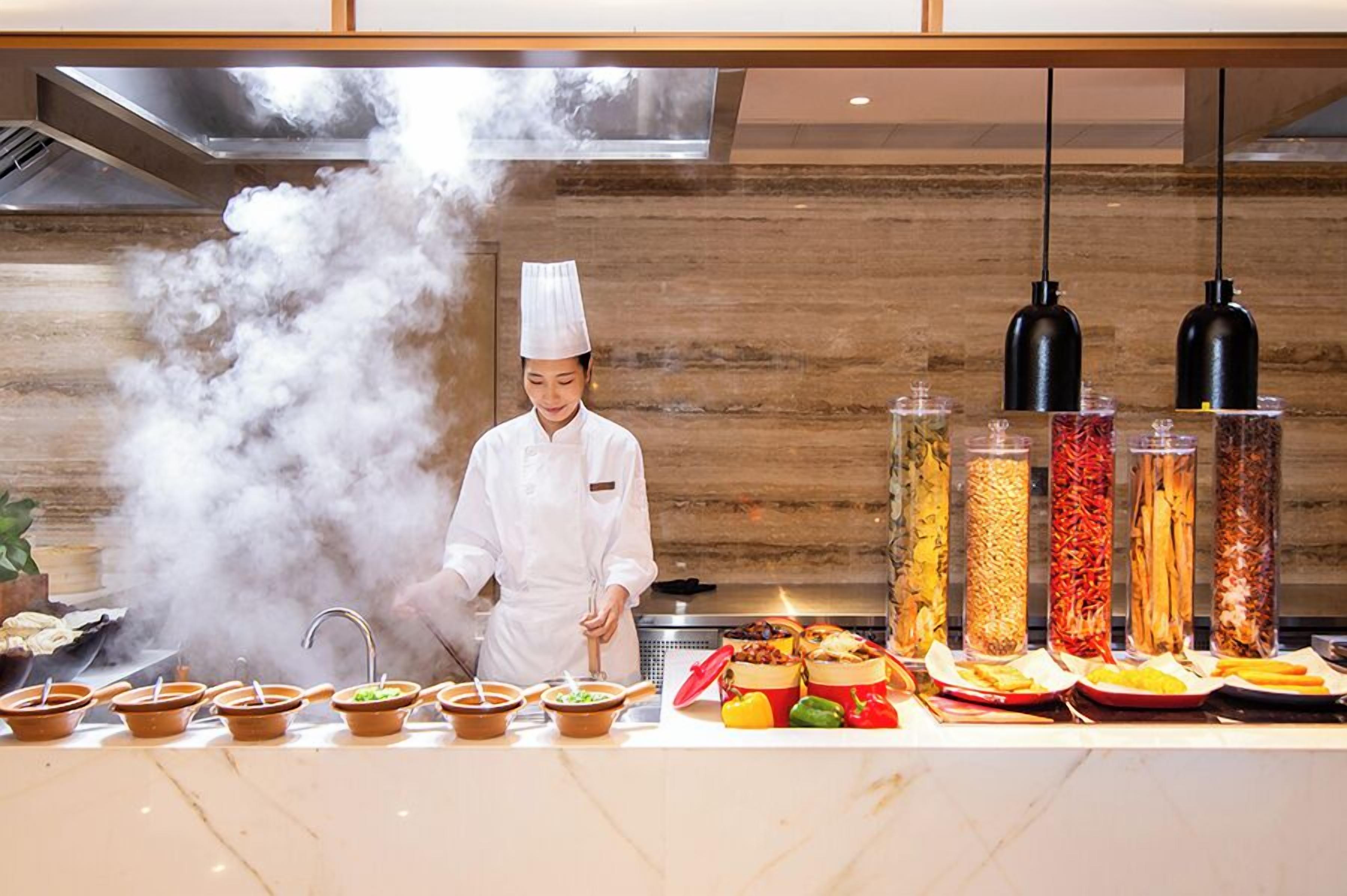 A chef in a white uniform cooking with steam rising in a modern kitchen with a wooden wall and hanging pendant lights.