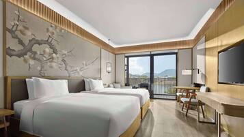 A hotel room with two beds, a desk, a chair, a TV, and a balcony with a view of the mountains.