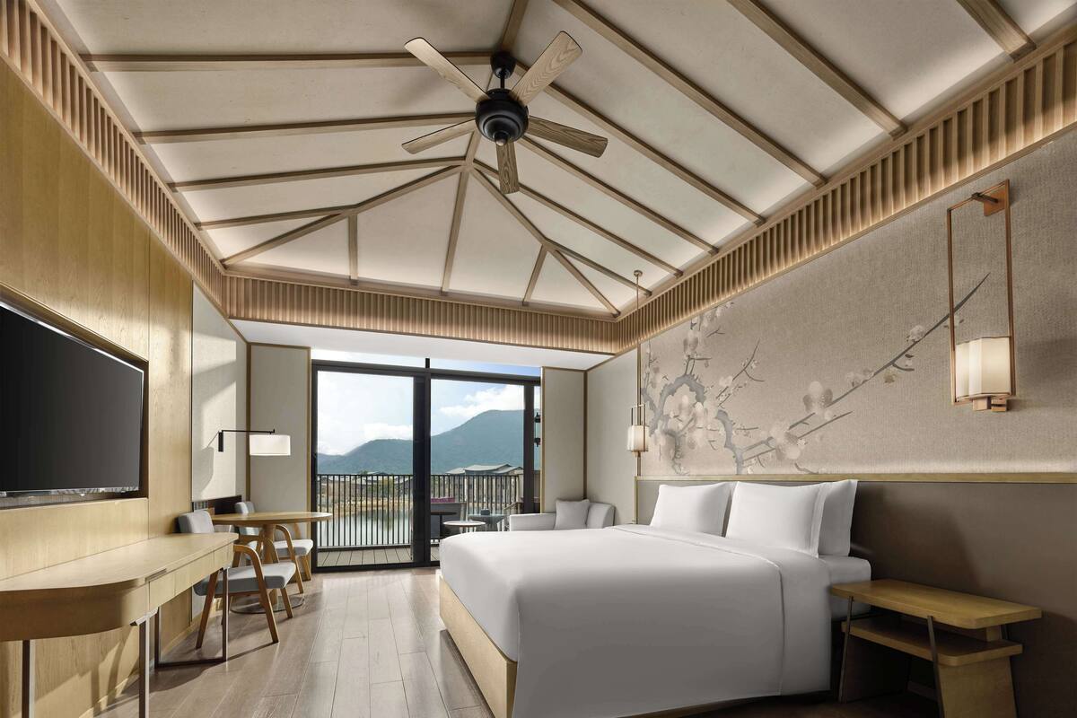 A hotel room with a large bed, a desk, and a TV. There is a balcony with a view of mountains.
