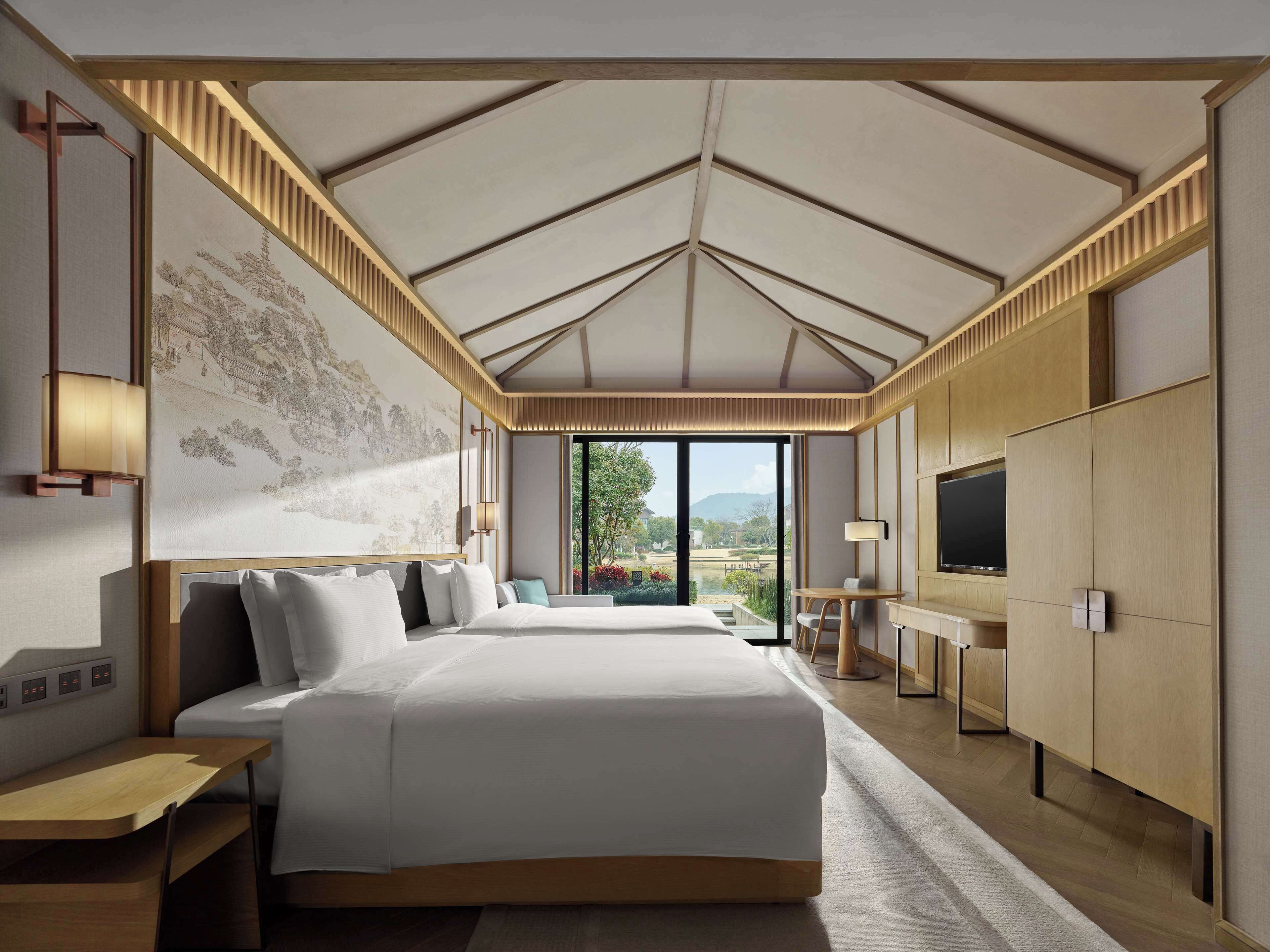 A spacious bedroom with a large bed, a desk, a TV, and a view of a garden.