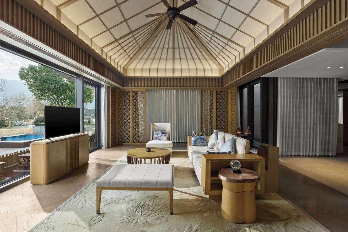 A spacious living room with a high ceiling, a ceiling fan, a large TV, a sofa, a coffee table, and a view of a swimming pool.