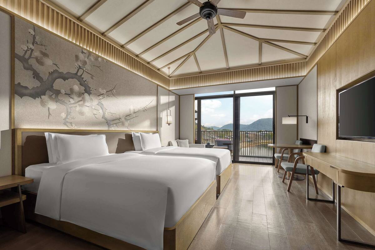 A modern hotel room with two beds, a desk, a balcony with a view, and a ceiling fan.