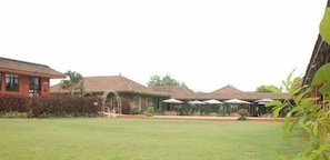 A courtyard with a lawn, a building with a red facade, and several thatched-roof structures with umbrellas. - The International Centre Goa (Panaji)
