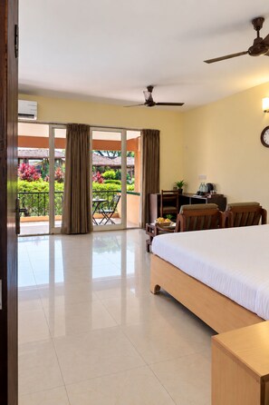 Quarto de hotel com cama, escrivaninha, cadeira, ventoinha no teto e vista para um jardim através de uma porta de vidro.