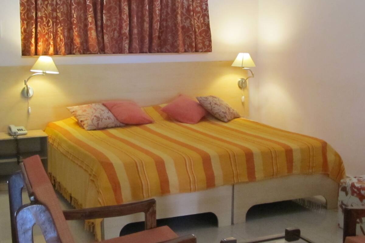 Habitación de hotel con cama, dos lámparas de noche, una silla, una mesita y un teléfono en la mesilla.