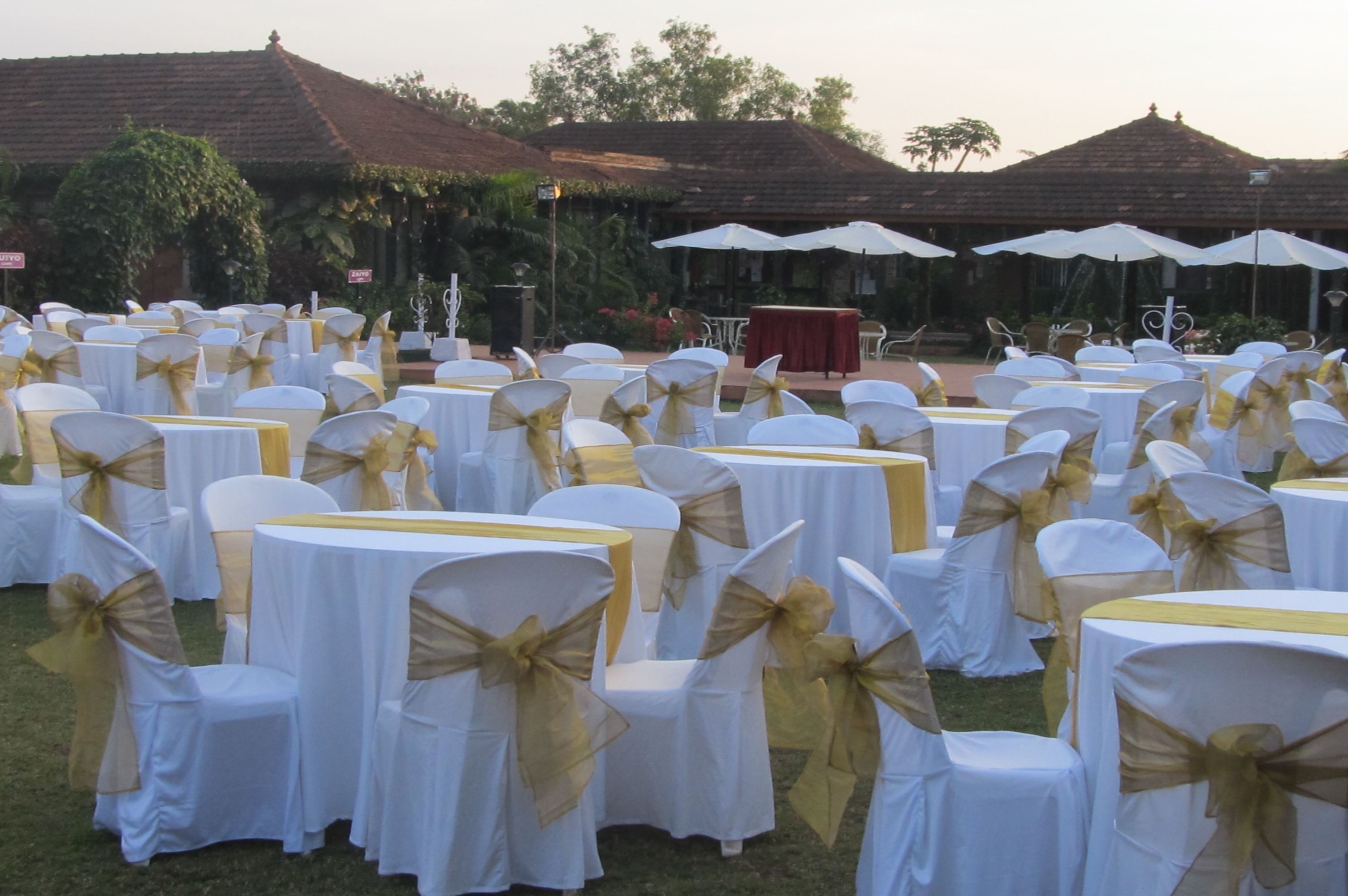 An outdoor event space with round tables covered in white tablecloths, chairs with gold sashes, and white umbrellas.