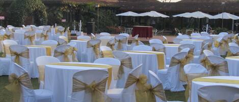An outdoor event space with round tables covered in white tablecloths, chairs with gold sashes, and white umbrellas.
