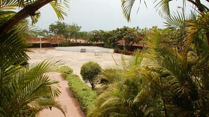 An outdoor patio area with a concrete floor, surrounded by lush greenery and palm trees. There are a few seating areas with benches and a table. - The International Centre Goa (Panaji)