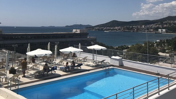 Una piscina en la azotea con sillones de descanso y sombrillas, con vistas a un paisaje urbano costero.