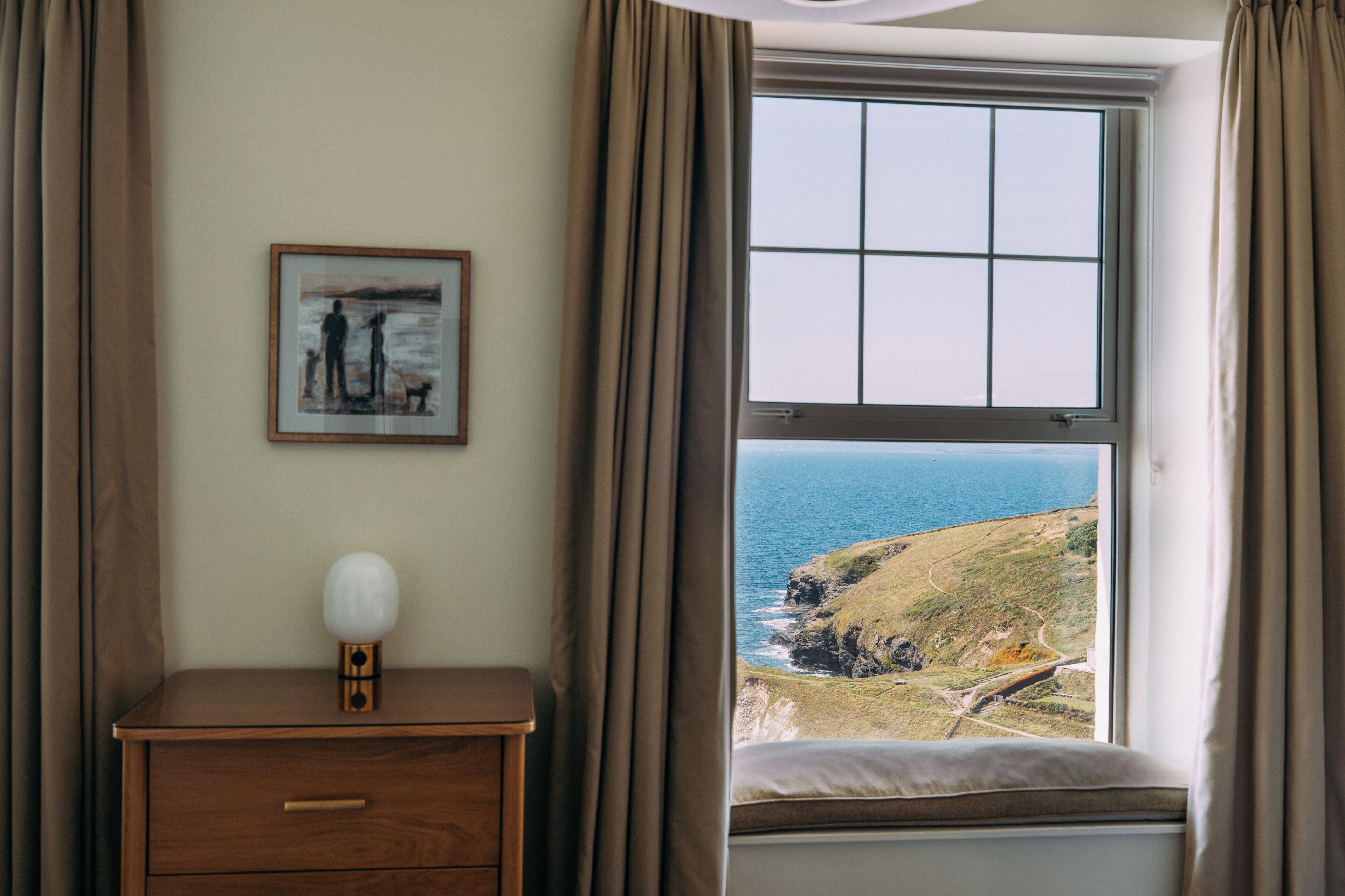 Habitación con un ventanal que ofrece vistas al mar, una mesita de noche con lámpara y un cuadro en la pared.