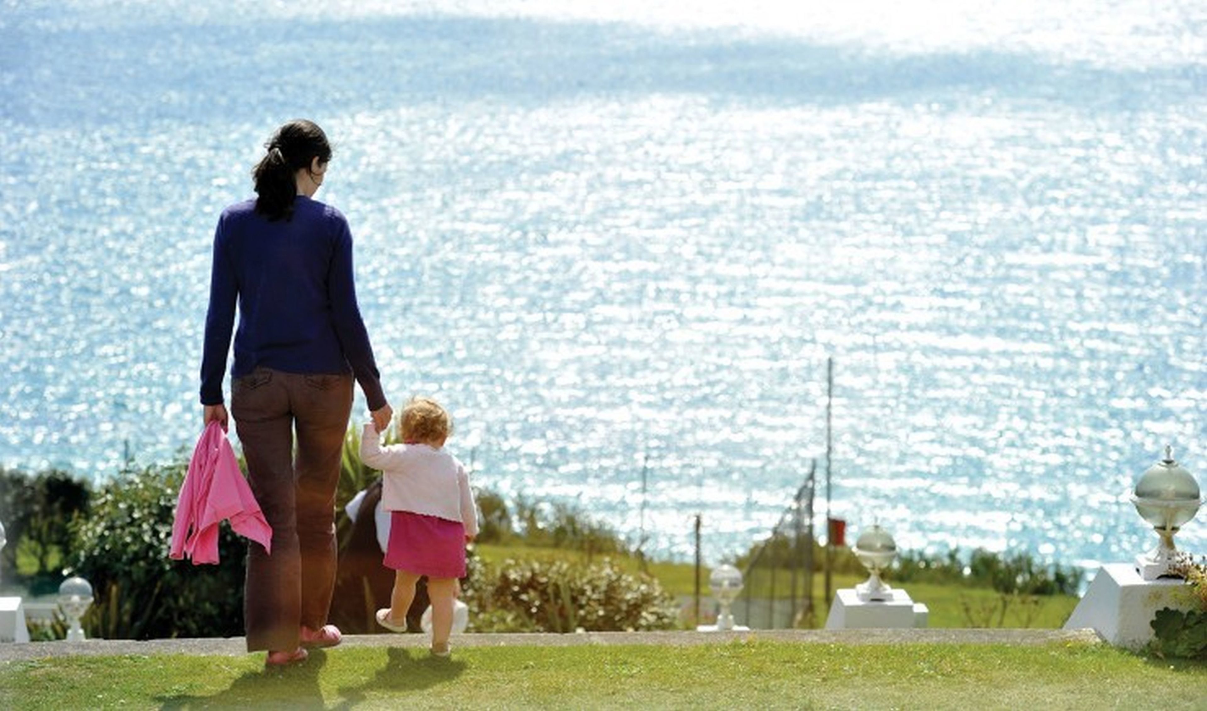 A woman and a child walking on a path with a view of a sparkling body of water.