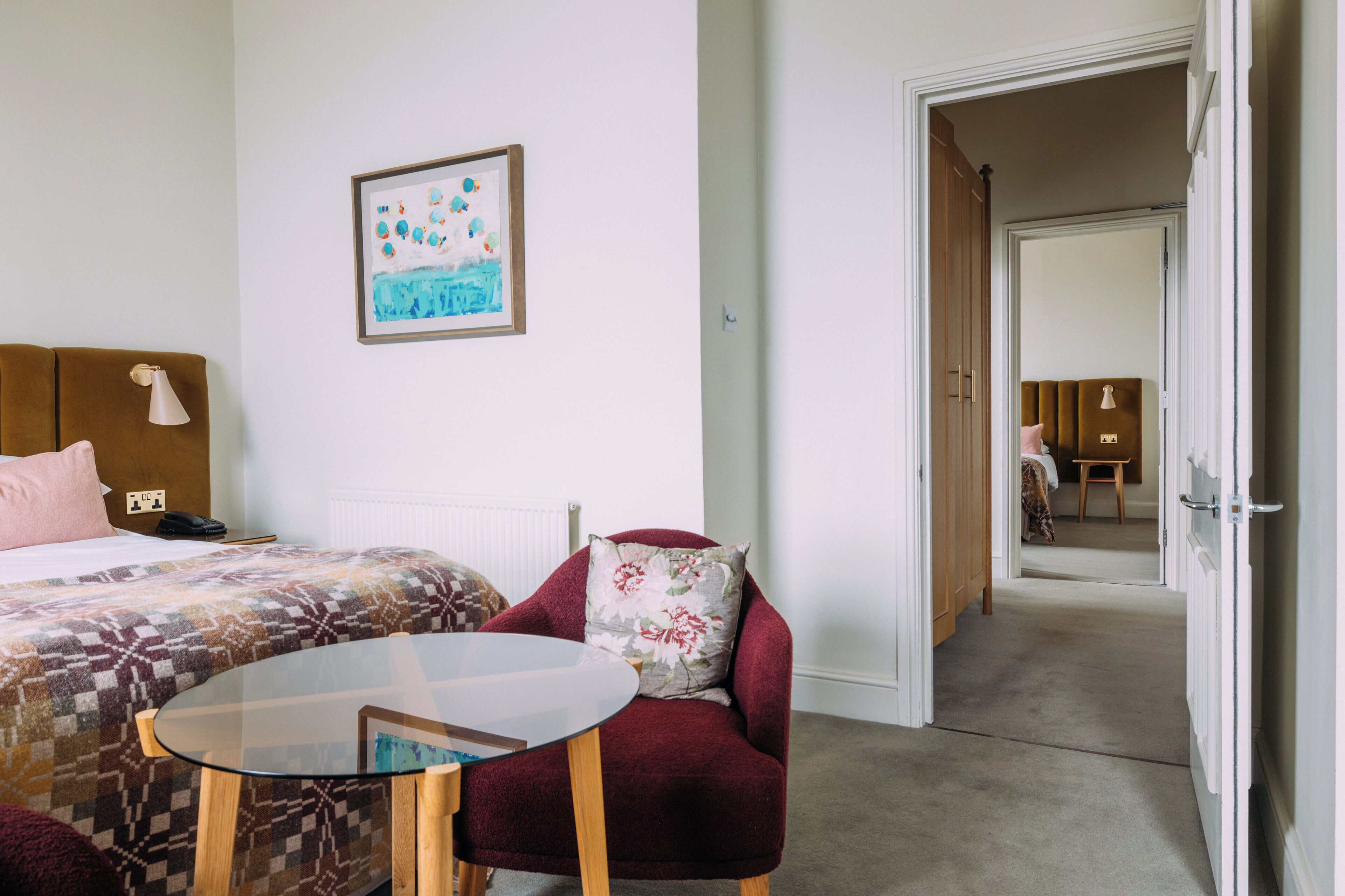 A hotel room with a bed, a chair, a table, and a painting on the wall.