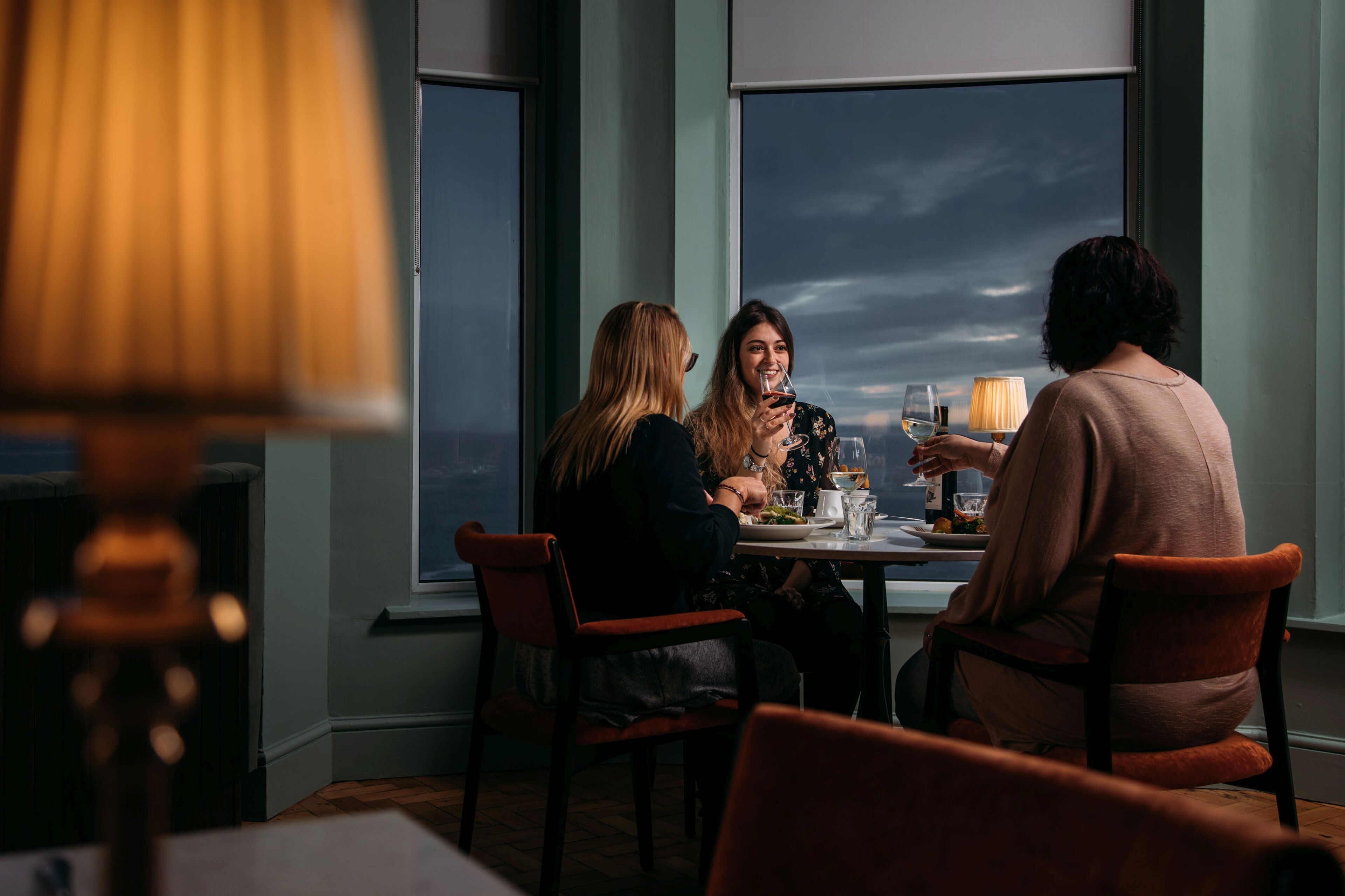 A dining area with a table set for three people, large windows, and a lamp on the table.