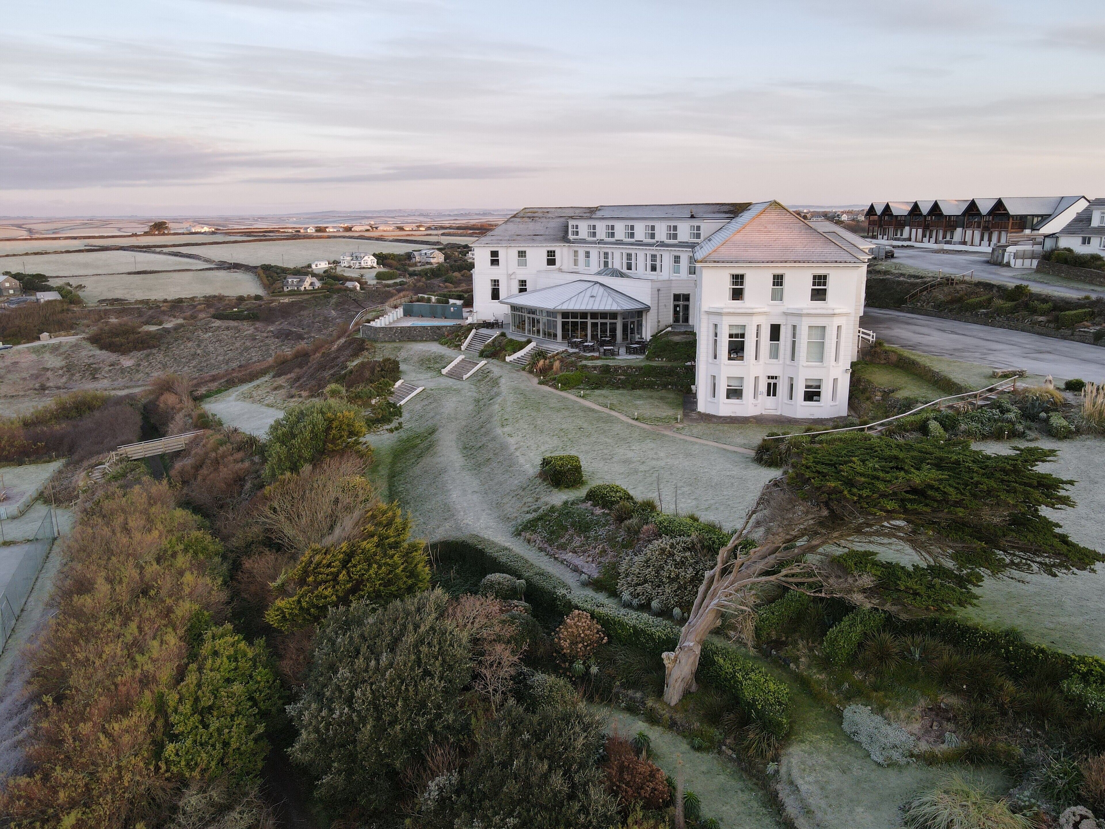Un grand bâtiment blanc, à étages multiples, surmonté d’une tour imposante, entouré de jardins aménagés et d’un paysage vallonné.