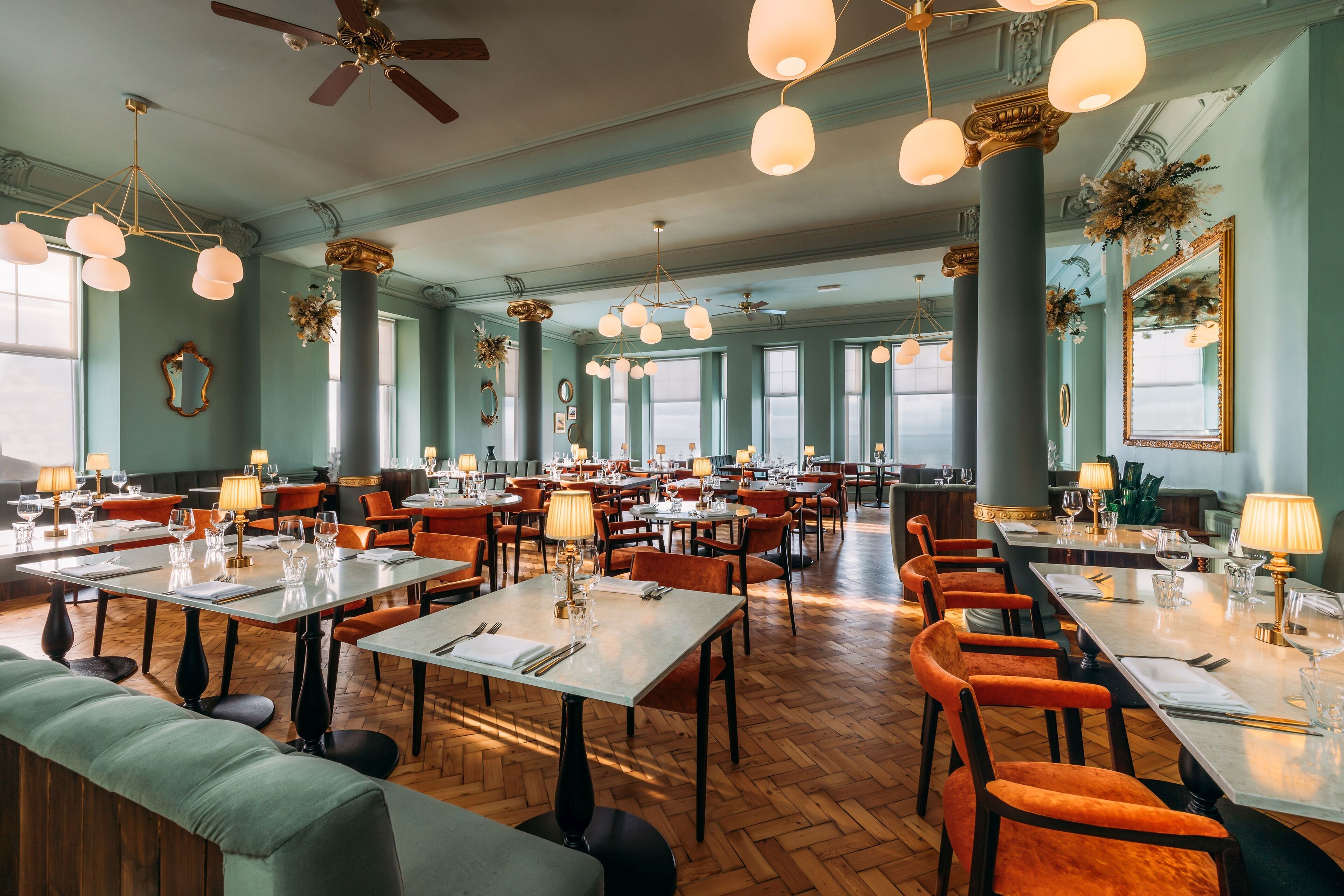 A spacious dining area with multiple tables and chairs, large windows, and decorative elements like flowers and mirrors.