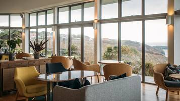 A spacious room with large windows offering a view of the hills. There are wicker chairs and a round table.