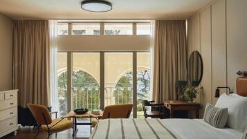 A hotel room with a large bed, a desk, two chairs, a dresser, and a balcony with a view.