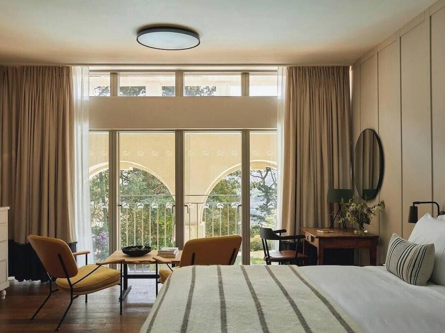 A hotel room with a large bed, a desk, two chairs, a dresser, and a balcony with a view.