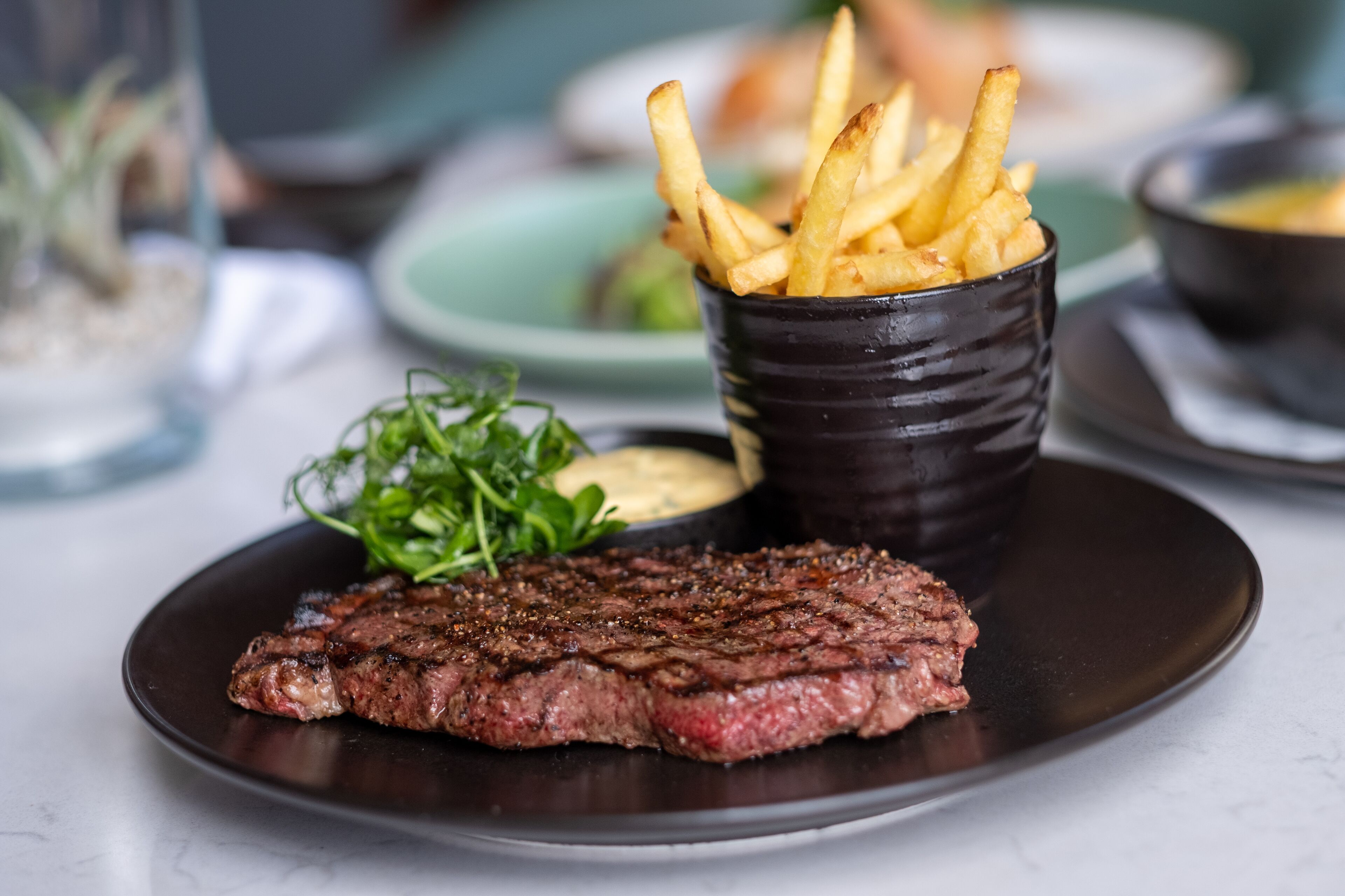 A plate with a steak and fries, a small bowl of sauce, and some greens on a table.
