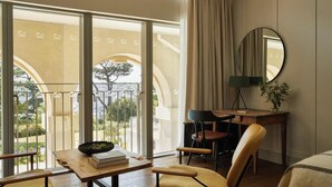 Ein Zimmer mit Schreibtisch aus Holz, zwei Stühlen, einem runden Spiegel und Blick auf einen Balkon mit Baum.