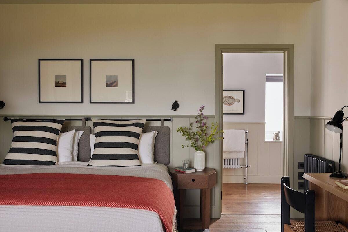 A bedroom with a bed, bedside table, and a desk with a chair. There are framed pictures on the wall and a radiator in the corner.