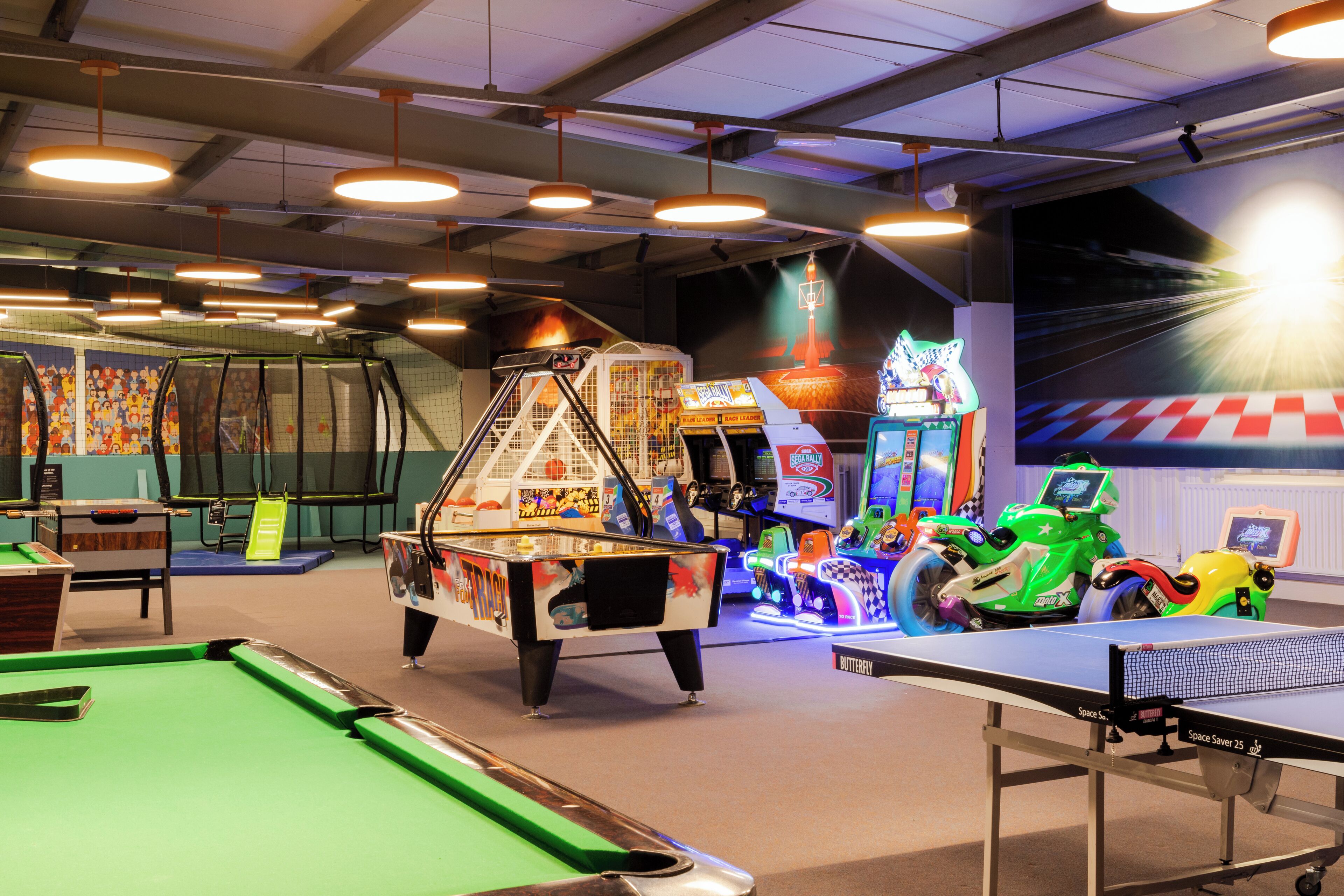 Un espace de loisirs comprenant une table de billard, un baby-foot, des jeux d’arcade et une fresque murale sur le thème de l’espace.