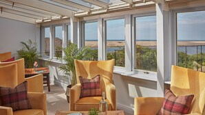 Una habitación soleada con amplios ventanales que dan a una playa, amueblada con sillones amarillos y una mesa de madera.