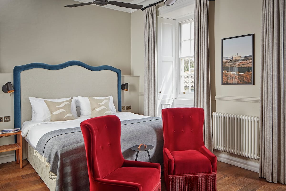 A bedroom with a large bed, two red chairs, a ceiling fan, a radiator, and a window with a view of a field.