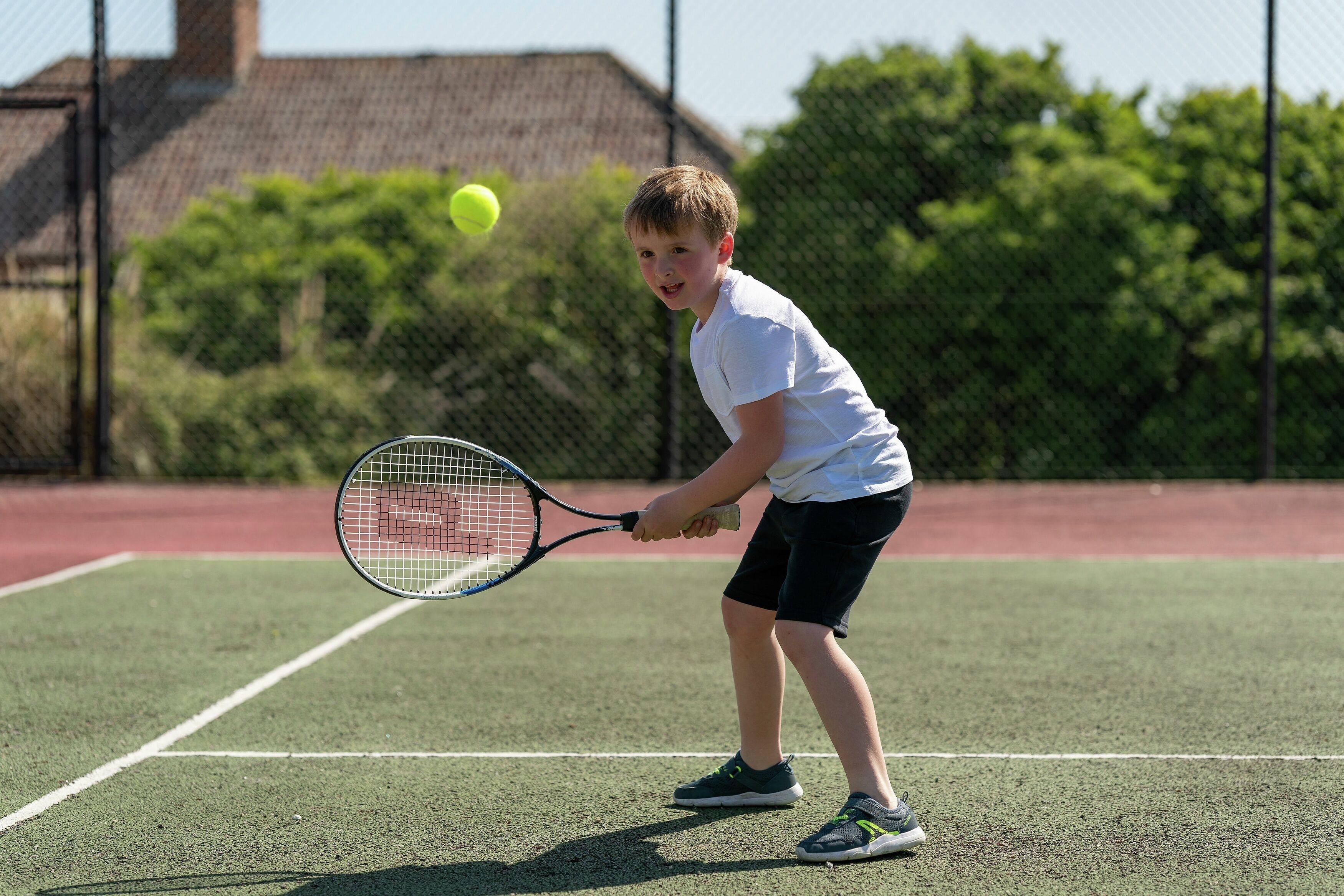 A tennis court with a player in a white shirt and black shorts holding a tennis racket, ready to hit a ball.