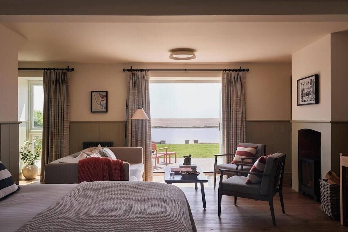 A hotel room with a large bed, a sofa, a chair, a small table, and a fireplace. There is a window with curtains and a view of the sea.