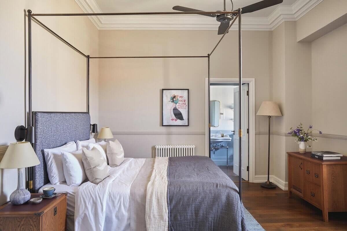 A bedroom with a four-poster bed, a ceiling fan, a radiator, a bedside table with a lamp, and a nightstand with a lamp.