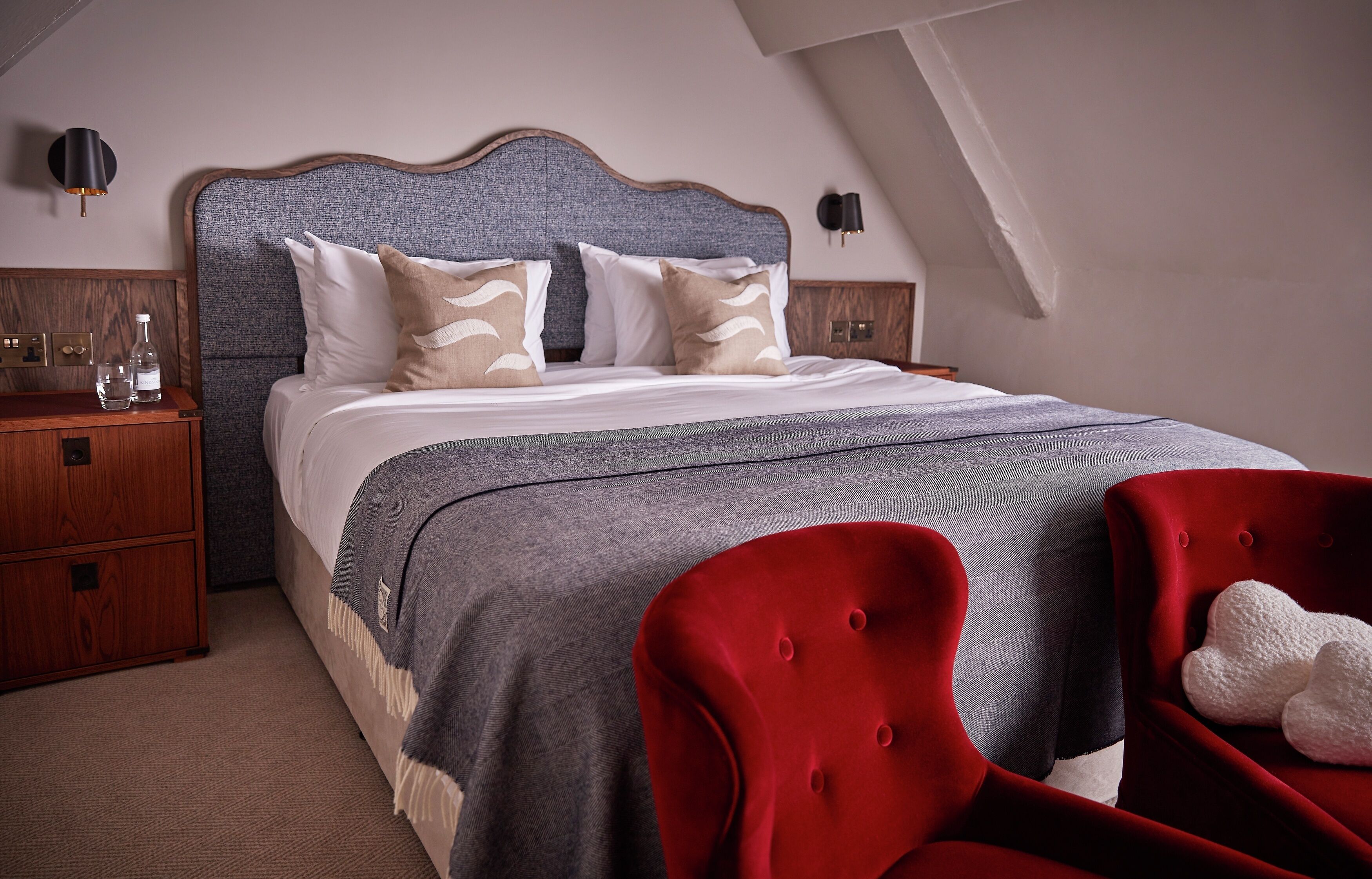 A bedroom with a large bed, a wooden nightstand, a red upholstered chair, and a wall-mounted lamp.
