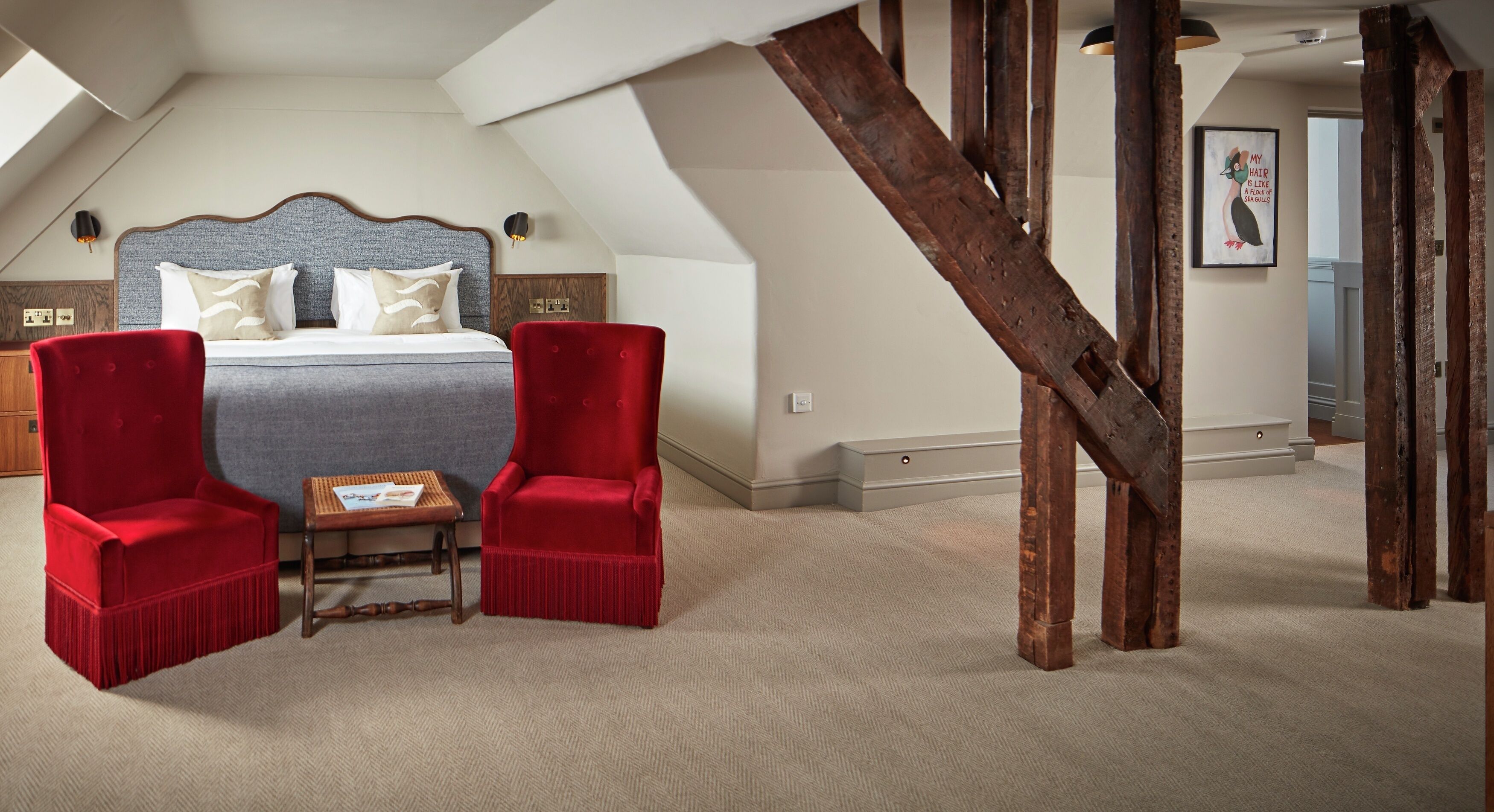 A bedroom with a large bed, two red chairs, a small table, and exposed wooden beams.