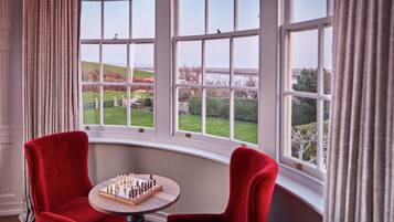 A room with a bay window, a red chair, a small table with a chessboard, and a view of a green landscape.