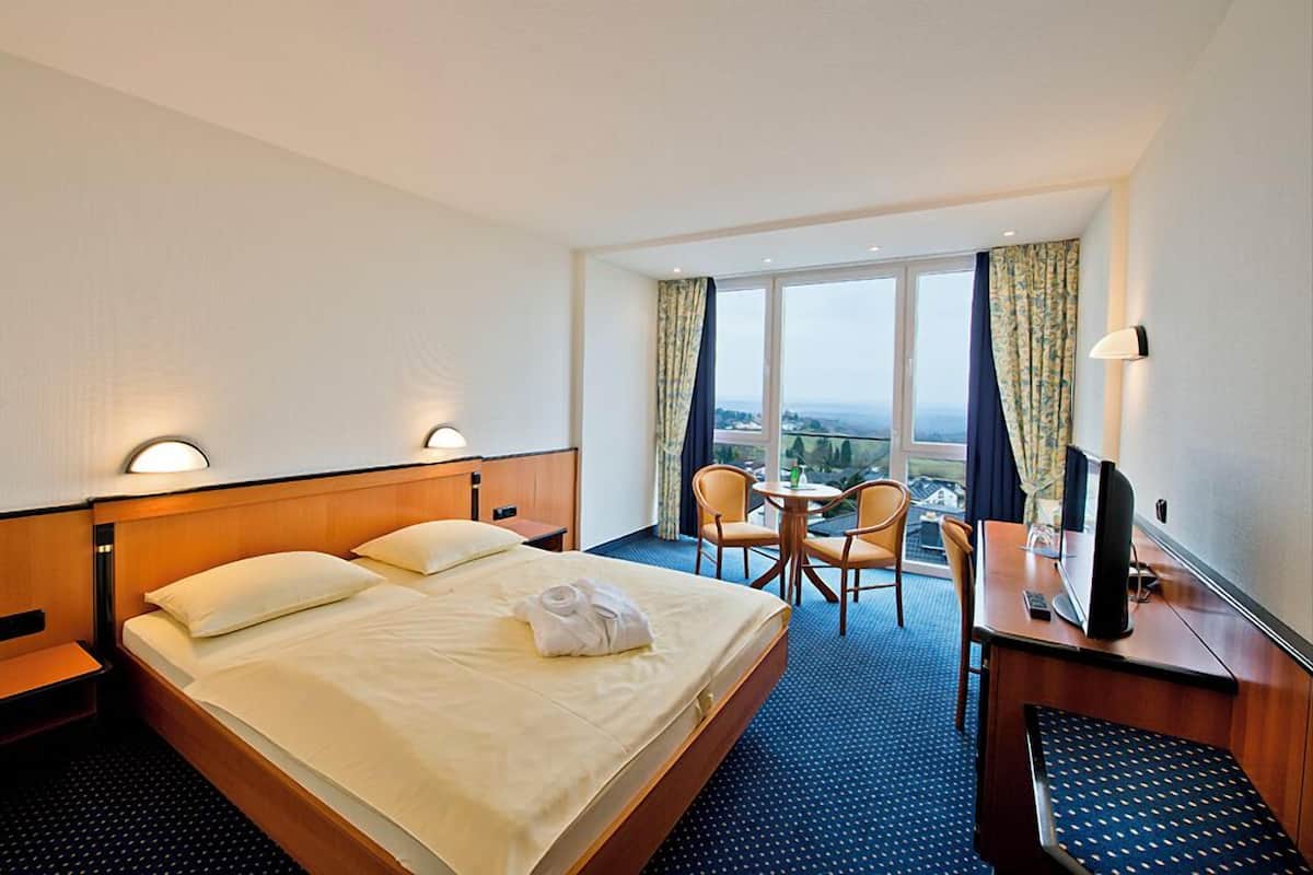 Camera d'albergo con un letto, due sedie, una scrivania con un televisore e un'ampia finestra con vista sulla città.