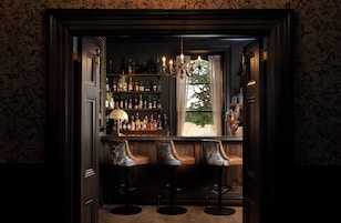 A bar area with a view of the outdoors, a chandelier, and a shelf of bottles.